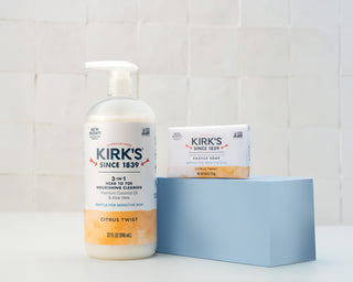 A bottle of Kirk’s 3-in-1 Nourishing Cleanser and a bar of Kirk’s Castile Soap sit on a light blue block against a white tiled background. Both products are labeled Citrus Twist and for sensitive skin.
