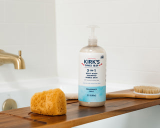 A bottle of Kirk’s 3-in-1 body wash, shampoo, and bubble bath sits on a wooden bath tray next to a yellow sponge and a brush, with a bathtub and gold faucet in the background.