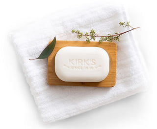 A white bar of soap labeled “Kirk’s Since 1839” rests on a wooden soap dish atop a folded white towel, with a small green branch and leaf placed beside it.